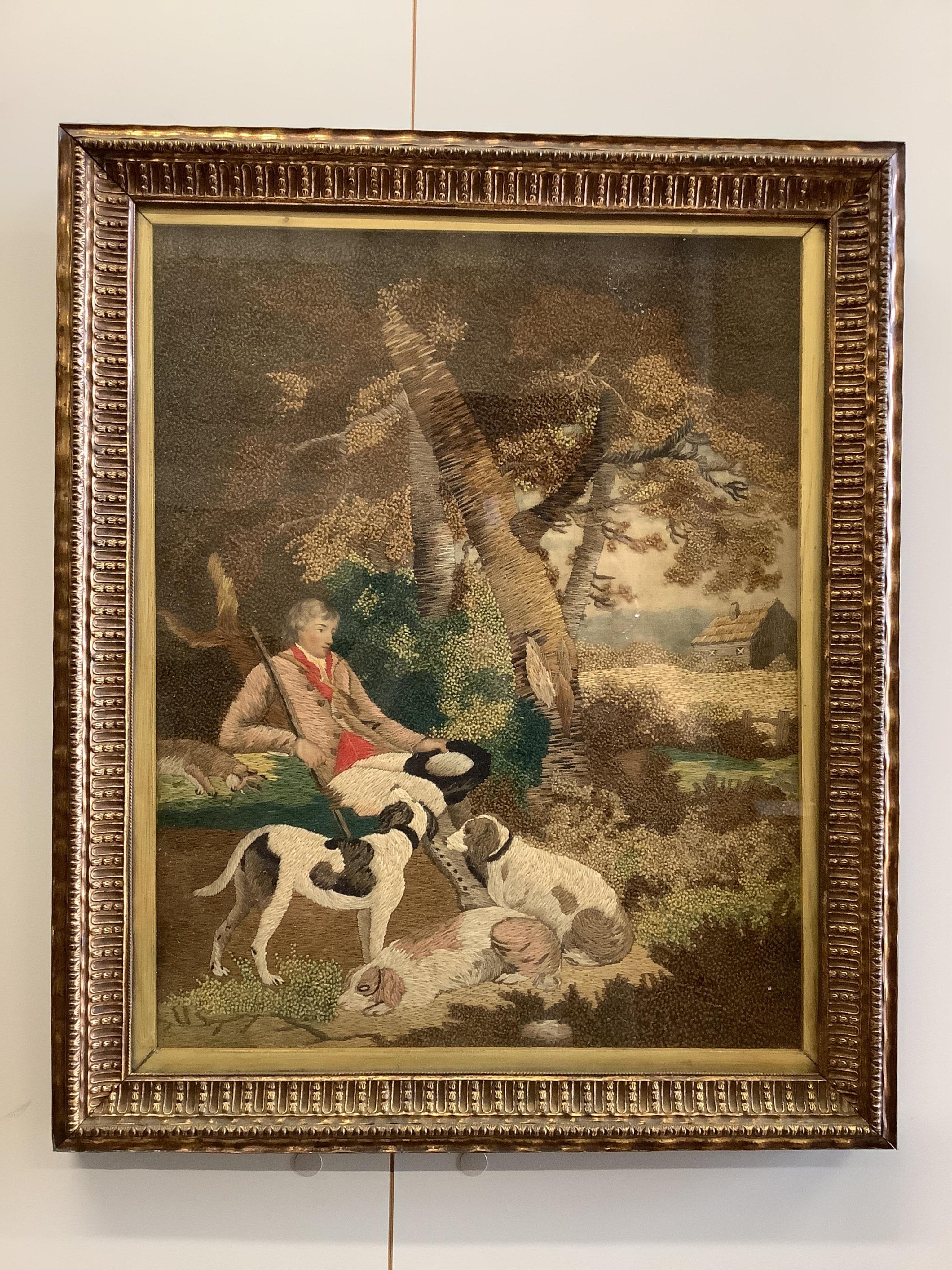 A framed early 19th century wool worked embroidery of huntsman sitting with a rabbit and his three hunting dogs under a tree, by a stream, with a field and cottage in the background, the embroidery worked in fine multi-c